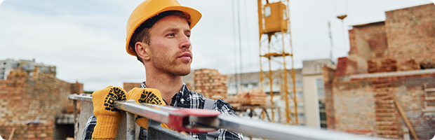 A construction worker on a job site wears safety gear and carries a ladder.  